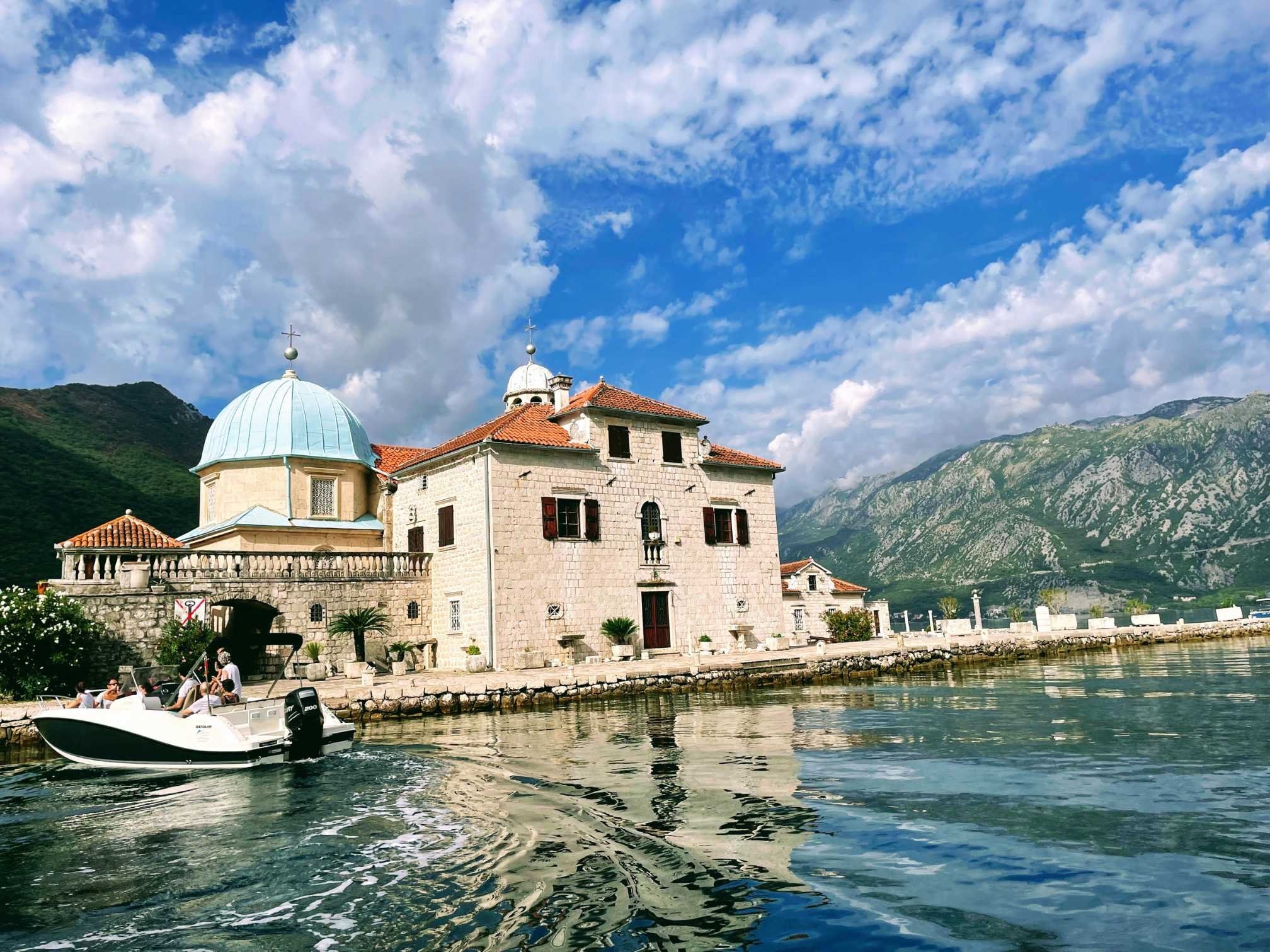 View of our Lady of the Rocks church surrounded by water, green mountains and and blue skies